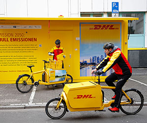 Zwei DHL-Boten mit einem DHL-Lastenfahrrad vor einer DHL-Station.