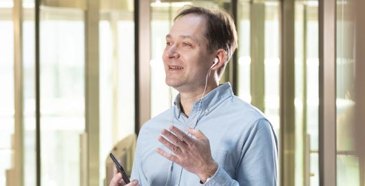 Bernd Neugebauer steht in der Empfangshalle des Bürogebäudes und telefoniert via Freisprechanalage und Kopfhörer mit seinem Smartphone. Er trägt ein hellblaues Hemd und eine beige Hose. Er gestikuliert.