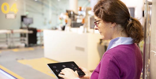 Alexandra Dietel steht im Büro und bedient ein Tablet, das sie in den Händen trägt. Sie trägt einen pinken Pullover und hat die Haare zusammengebunden.