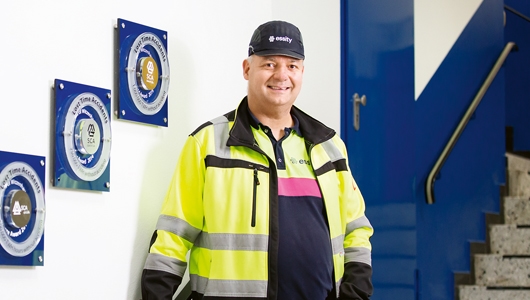 Porträt von Bernd Witzke, Fachkraft für Arbeitssicherheit bei Essity am Standort Witzenhausen. Er trägt einen schwarzen Schutzhelm, Warnkleidung mit waagerechten und senkrechten Streifen sowie ein Poloshirt, auf dem der Unternehmensname steht.