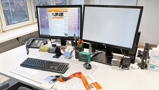 Ein Bildschirmarbeitsplatz der Berliner Stadtreinigung, Auf dem Schreibtisch stehen zwei große Computerbildschirme, eine schwarze Tastatur, Telefon, Maus und weitere Schreibtischutensilien. Ein Schlüsselbund mit orangenem Umhängeband liegt ebenfalls auf dem Schreibtisch.