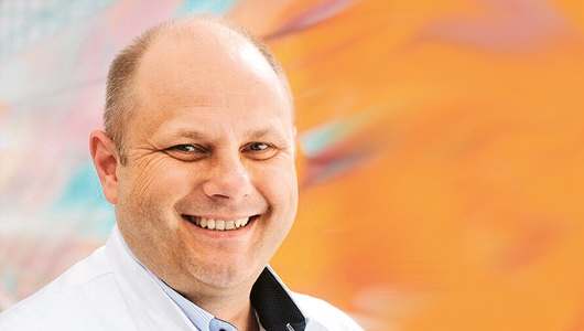 Porträt von Dr. Ingolf Hosbach, stellvertretender Leiter der Poliklink des Instituts für Prävention und Arbeitsmedizin der Deutschen Gesetzlichen Unfallversicherung (IPA). Er hat eine Halbglatze, ein weißes Hemd mit offenem Kragen an und lächelt.