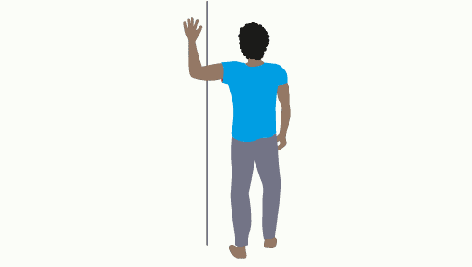 Grafik einer Person mit schwarzen, gelockten kurzen Haaren. Sie trägt ein blaues T-Shirt und eine graue Hose. Die Person hält den linken Arm im rechten Winkel gegen eine Ecke einer Wand. Der rechte Arm liegt am Körper an. Der rechte Fuß ist leicht nach vorn gestellt.