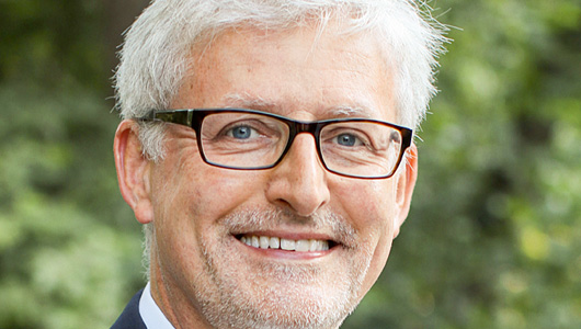 Ein Porträt von Uwe Marx. Er trägt eine eckige Brille, ein Hemd und Sakko und lächelt in die Kamera. Er hat kurzes, weißes Haar. Im Hintergrund sind Bäume zu sehen.
