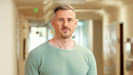 Porträt von Pflegedienstleiter Mathias Kaupp. Er schaut frontal in die Kamera und lächelt ganz leicht. Die blond-grauen Haare trägt er kurz und er hat einen braunen Drei-Tage-Bart. Er trägt einen graugrünen Pullover.