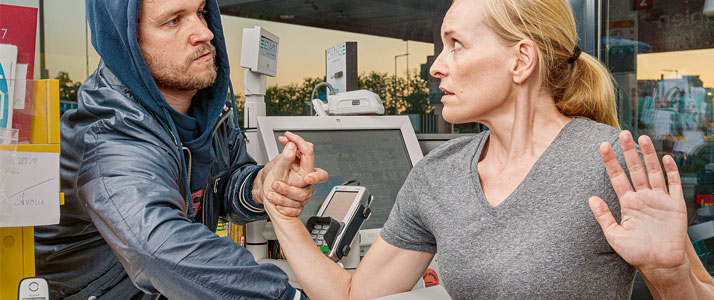 Ein Mann in Kapuze und Lederjacke greift mit seiner Hand über einen Tresen hinweg in eine geöffnete Kasse. Er schaut dabei grimmig zur Frau an der Kasse. Mit der anderen Hand hält er gewaltvoll eine Hand der Frau fest. Sie hat beide Arme erhoben und blickt ängstlich zum Räuber.