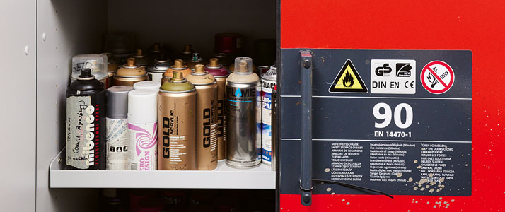 Blick in einen geöffneten roten Gefahrstoffschrank. Im Schrank stehen auf einem weißen Regalbrett verschiedene bunte Farbflaschen.