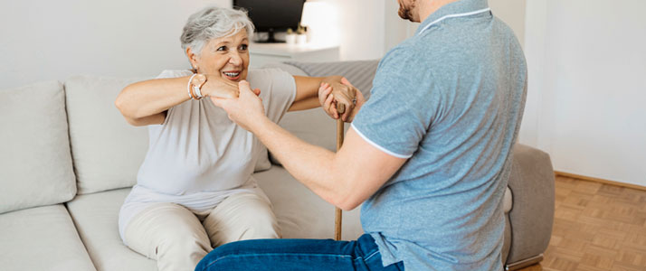 Eine Seniorin sitzt auf einem Sofa und ist im Begriff aufzustehen. In der einen Hand hält sie einen Stock zum Abstützen und die andere Hand wird von einer Pflegekraft gehalten, die mit geradem Rücken vor der Seniorin sitzt.