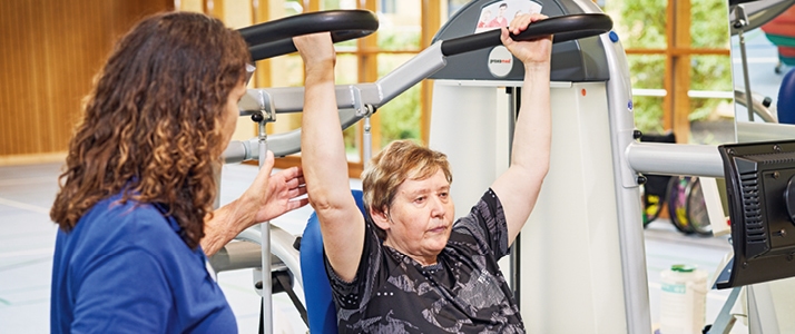 Altenpflegerein Inka Bienek sitzt auf einem Fitnessgerät und hat die Arme über den Kopf gehoben. Links von ihr steht eine Therapeutin, die die Übung anleitet.