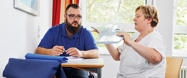 Arbeitstherapeut Marcus Günther sitzt mit der Altenpflegerin Inka Bienek an einem Tisch. Inka Bienek betrachtet ein Rutschbrett.