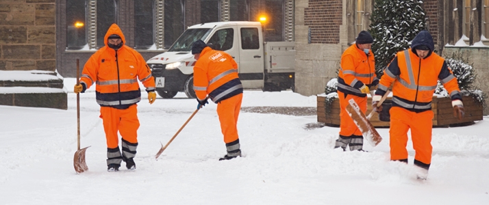 Vier Beschäftigte vom Winterdienst schippen Schnee in einer Straße. Sie tragen Warnkleidung in Form von Warnjacke- und Hose. Zudem tragen Sie dicke Wollmützen.