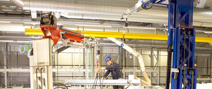 In einer Werkshalle der Firma Multivac wechselt Sascha Ostrowski aus dem Team Instandhaltung ein Kühlaggregat aus. Er befindet sich auf dem Dach einer großen Produktionsanlage. Am Boden davor steht ein Kollege, der einen mobilen Kran steuert, an dem das Kühlaggregat befestigt ist. Der Kollege auf dem Dach der Maschine trägt eine persönliche Schutzausrüstung gegen Absturz, die an einem mobilen Anschlagpunkt befestigt ist.