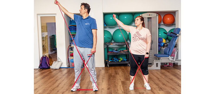 Simone Lorenz und Physiotherapeut Nazif Aktas trainieren in der Physio-Praxis nebeneinander mit elastischen Bändern. Mit einem Arm ziehen sie das Band nach oben, die Füße stehen auf den Bändern.