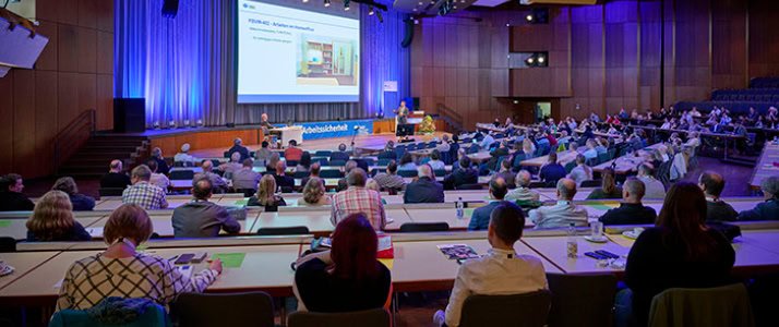 Ein größer Hörsaal mit Holzvertäfelung und vollbesetzten Sitzreihen. Auf einem Podium referiert ein Mann, an der Rückwand ist eine große Leinwand angebracht, auf der eine Powerpoint Präsentation zu sehen ist.