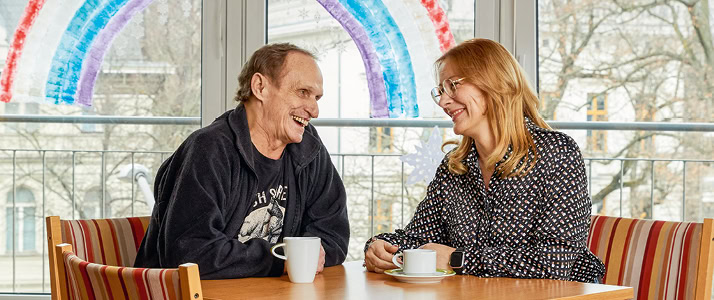 Heimbewohner Michael Keßner und Heimleiterin Nicole Oerder sitzen an einem Tisch im Aufenthaltsraums des Immanuel Seniorenzentrums Schöneberg. Sie schauen sich an und lachen. Im Hintergrund ist ein großer Regenbogen an die Scheibe gemalt. Michael Keßner hat braunes, kurzes Haar und einen schwarzen Pullover, Nicole Oerder trägt langes, blondes Haar und eine gemusterte Bluse.
