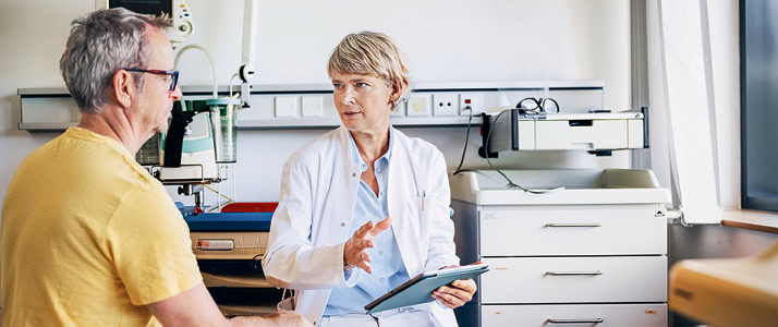 Eine Betriebsärztin bespricht etwas mit einem Patienten. Sie trägt einen weißen Kittel, eine weiße Hose und ein blaues Hemd. Der Patient trägt ein gelbes Shirt, eine graue Hose und eine Brille.