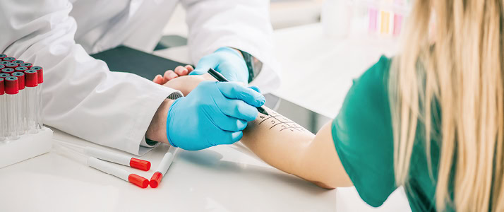 Szene in einer ärztlichen Praxis. Abgebildet sind die Arme einer Person in weißem Kittel und blauen Latexhandschuhen, die mit einem Stift ein Raster für einen Allergietest auf den Arm einer anderen Person malt. Diese Person trägt ein grünes Shirt.