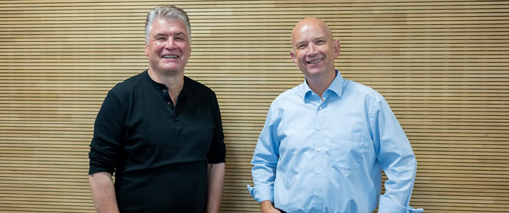 Thomas Assmann und Steffen Stech von der Still GmbH stehen nebeneinander und lachen in die Kamera. Assmann trägt ein schwarzes Shirt und hat graue, kurze Haare, Stech trägt ein hellblaues Hemd und hat eine Glatze. Sie stehen vor einer Holzwand.