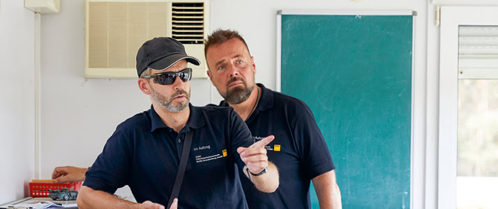 Stefan Prasdorf und René Payard vom Fahrsicherheitszentrum Berlin-Brandenburg des ADAC stehen gemeinsam. Sie tragen Polo-Hemden mit dem ADAC-Logo, Payard trägt Kappe und Sonnenbrille und gestikuliert.