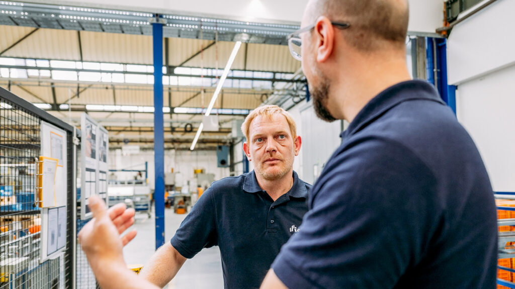 Nils Ludwig und Kolja Gowert stehen sich gegenüber und sprechen über Ergonomie. Beide haben kurze Haare und tragen dunkelblaue Poloshirts.