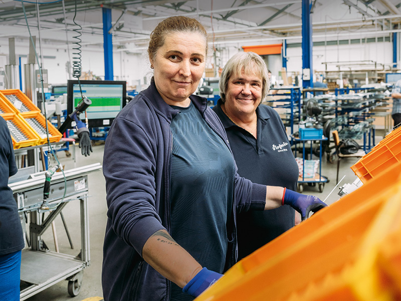 Zwei Frauen stehen in einer Fabrikhalle an einem Arbeitstisch. Beide schaue lächelnd in die Kamera.