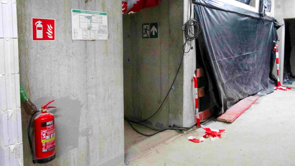 Rohbau mit montiertem Feuerlöscher an Betonstütze und einer Plane an der Wand.
