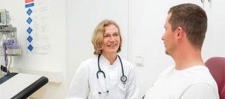 Eine Ärztin mit Stethoskop um den Hals im Gespräch mit einem Patienten, der auf einem Behandlungsstuhl sitzt.