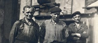Historisches Schwarz-Weiß-Bild dreier nebeneinanderstehender Männer. Der Mann in der Mitte träge eine Schiebermütze, der Mann rechts verschränkt die Arme vor der Brust und trägt ebenfalls eine Schiebermütze.