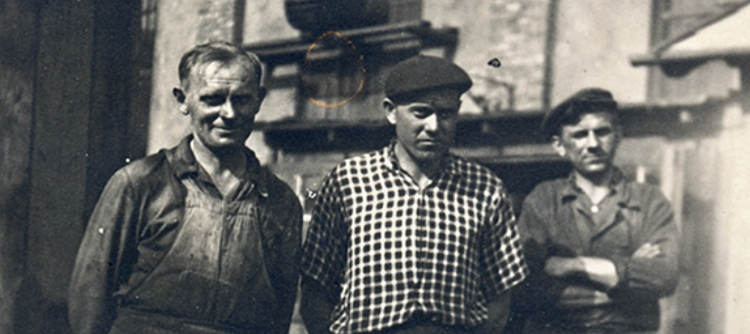 Historisches Schwarz-Weiß-Bild dreier nebeneinanderstehender Männer. Der Mann in der Mitte träge eine Schiebermütze, der Mann rechts verschränkt die Arme vor der Brust und trägt ebenfalls eine Schiebermütze.