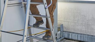 Eine Person mit hellbrauner Arbeitshose und Sicherheitsschuhen steigt eine Aluminiumleiternach oben. Die Situation findet draußen statt.