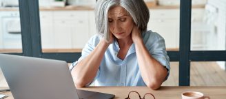 Eine Frau mit grauen Haaren sitzt vor einem Laptop am Tisch. Sie hat ihre Brille abgenommen, runzelt die Stirn und greift mit den Händen in ihren Nacken.
