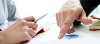 Jeweils eine Hand von zwei einander an einem Tisch gegenübersitzenden Personen zu sehen. Person linsk hält einen Kugelschreiber in der Hand, die Person rechts zeigt auf ein auf dem Tisch liegendes Dokument mit Diagrammen.