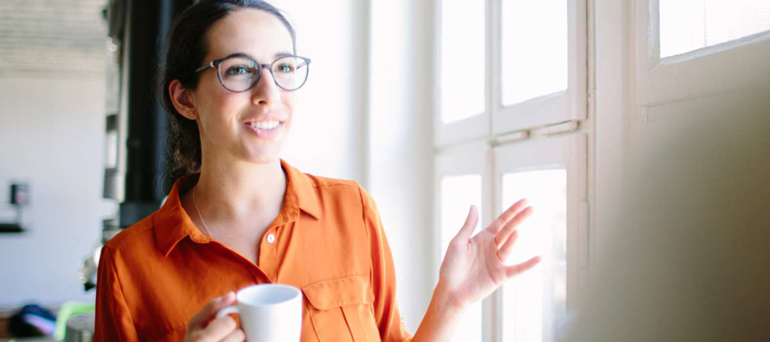 Eine Frau in orangener Bluse hält eine Kaffeetasse in der Hand und gestikuliert fröhlich beim Sprechen.