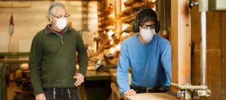 Zwei Männer stehen in einer Holzwerkstatt an der Werkbank. Einer von arbeitet mit einer Holzfläche, trägt einen Hör- und Atemschutz.