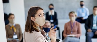 Eine weibliche Person steht im Vordergrund vor einem Publikum und hält einen Vortrag. Sie deutet mit einem Zeigeobjekt auf die Vortragsfläche und trägt eine Mund-Nase-Bedeckung.