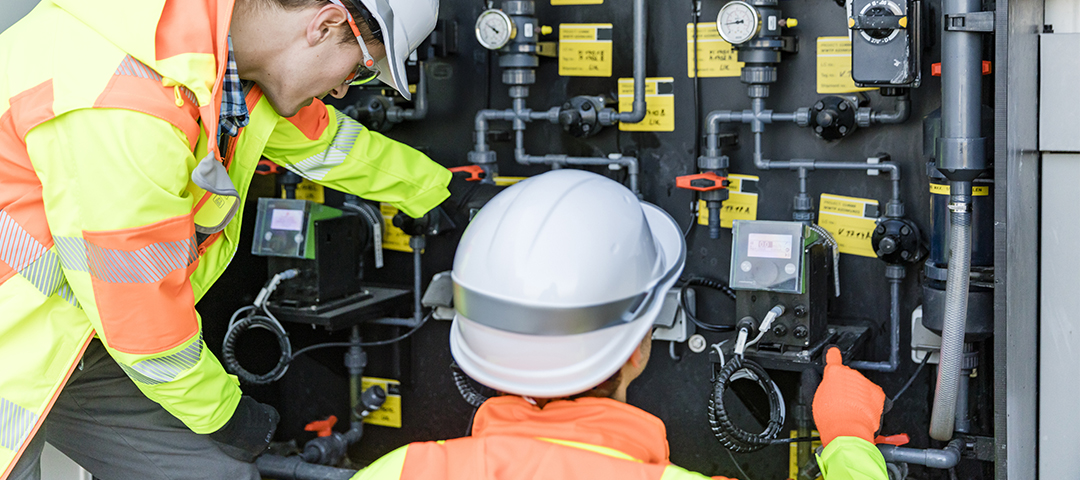 Zwei Mitarbeitende prüfen die Rohrleitungen an einer Maschine. Die beiden Personen tragen weiße Schutzhelme, Schutzbrillen, Schutzhandschuhe sowie gelb-orangene Schutzjacken.