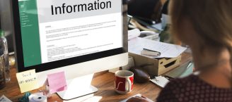 Eine Person sitzt an ihrem Arbeitsplatz an ihrem Schreibtisch vor ihrem Computer. Auf dem Computerbildschirm ist groß die Aufschrift "Information" zu sehen.