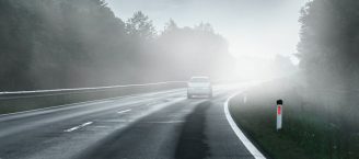 Auf einer Landstraße fährt im Halbdunkeln in einer Kurve ein einzelnes Auto. Die Sicht ist aufgrund von Nebel sehr schlecht.