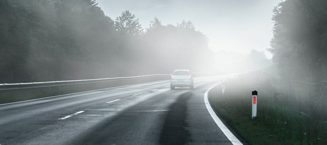Auf einer Landstraße fährt im Halbdunkeln in einer Kurve ein einzelnes Auto. Die Sicht ist aufgrund von Nebel sehr schlecht.