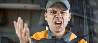 Ein Mann am Steuer eines Transporters ist wütend. Er zeigt mit einer Hand drohend zur vorausfahrenden Person, hat den Mund geöffnet und runzelt grimmig die Stirn.