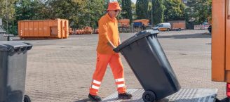Ein Müllwerker in langer Kleidung schiebt auf einem Betriebhof eine Tonne zum Müllwagen.