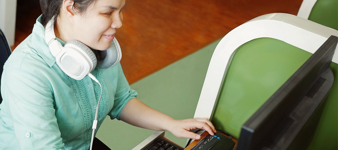 Person mit Down-Syndrom sitzt an einem Schreibtisch und ist am PC am arbeiten. Sie trägt ein grünes T-Shirt, kurzes, braunes Haar und weiße On-Ear Kopfhörer.