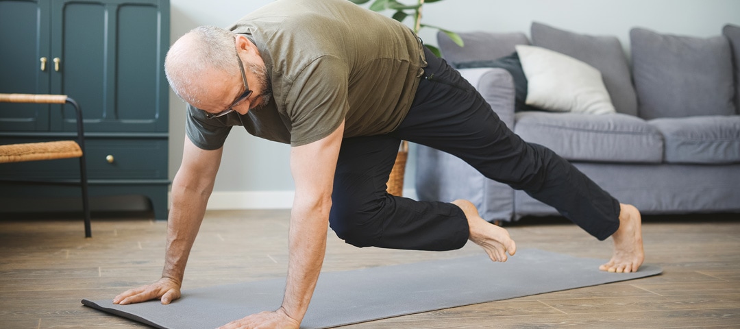 Ein Mann mit einem kakifarbenen Shirt und einer dunklen Jogginghose macht eine Sportübung auf einer hellgrauen Yogamatte. Er trägt eine Brille und hat rasierte Haare. Er befindet sich in einem Wohnzimmer.