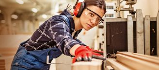 Eine Frau in Blaumann, einem karierten Hemd, roten Handschuhen und roten Kopfhörern. Sie schneidet konzentriert ein Holzbrett zurecht. Die Frau befindet sich in einer Werkstadt.