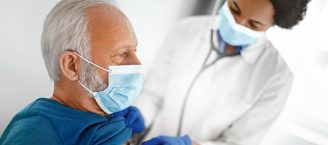 Ein Mann mit grauem Haar und einer Mundschutzmaske wird von einer Ärztin abgehört. Sie hält das Stethoskop an seine Brust. Die Ärztin trägt einen weißen Kittel.