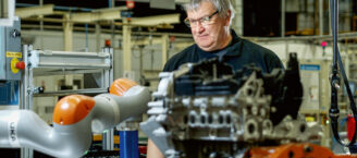 Dietmar Brauner, Produktionsmitarbeiter bei Ford, steht an einer Maschine in einer Produktionshalle. Er hat graue, kurze Haare und trägt eine randlose Brille und ein schwarzes Shirt. Sein Blick geht nach links unten auf die Maschinen. Links neben ihm ragt ein grau-orangefarbener Roboterarm ins Bild.
