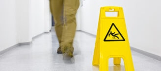 Eine Person in khakifarbener Hose läuft durch einen weißen Gang, nur Füße und Beine sind zu sehen. Vor ihr steht ein gelbes Warnschild auf dem Boden, das vor rutschigem Boden warnt. Ein schwarzes Symbol auf dem Schild zeigt eine ausrutschende Figur in einem Dreieck.