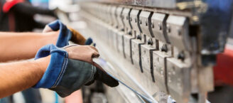 Eine Person arbeitet an einer Maschine, die Metall biegt. Ihre Hände sind durch Schutzhandschuhe geschützt.