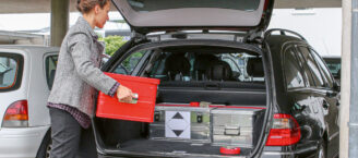 Eine Frau lädt eine rote Kiste in den geöffneten Kofferraum eines Autos. Im Kofferraum stehen bereits zwei Kisten aus Metall. Ein Aufkleber zeigt, dass es sich um Gefahrgut handelt.