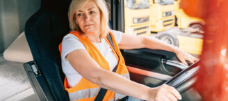 Eine Frau im mittleren Alter rangiert mit einem Lkw und schaut dabei in einen Seitenspiegel. Sie trägt eine orangene Warnweste über ihrem weißen-T-Shirt. Sie ist angeschnallt.